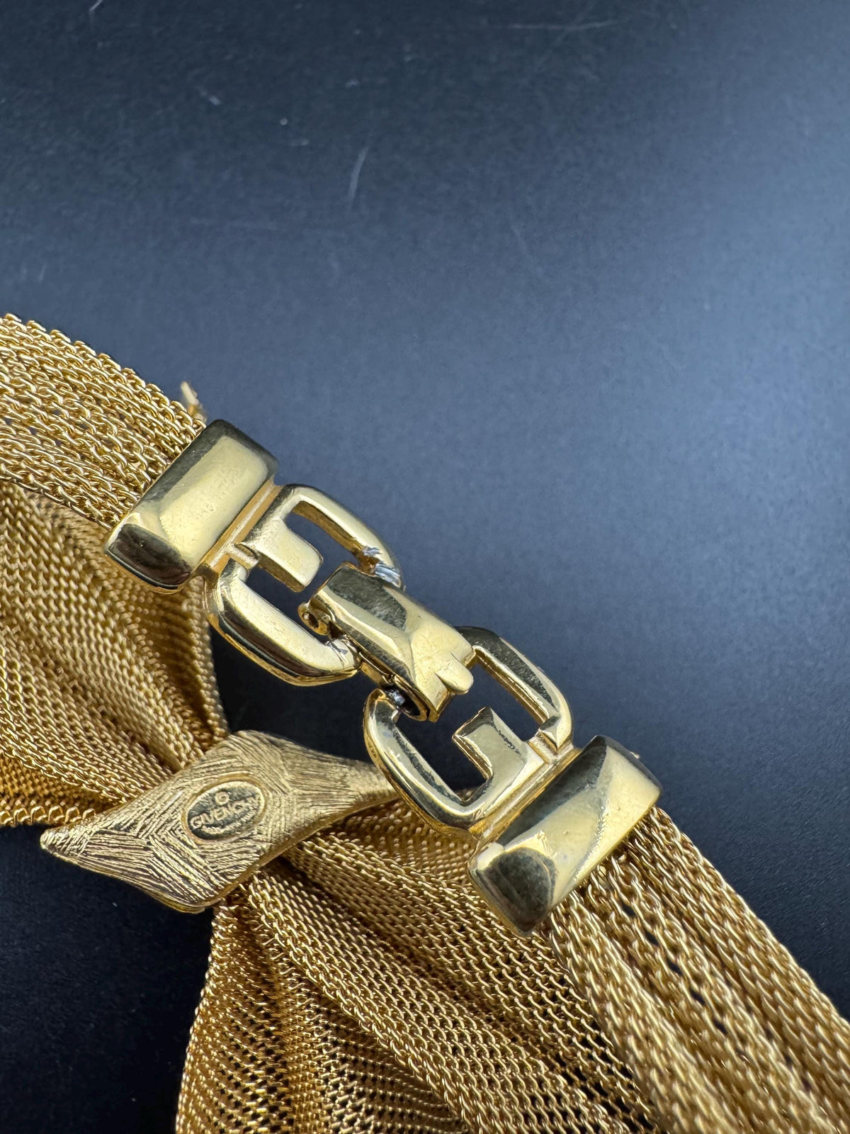 A close-up view of a gold-colored metal mesh bracelet with a clasp and a decorative element.
