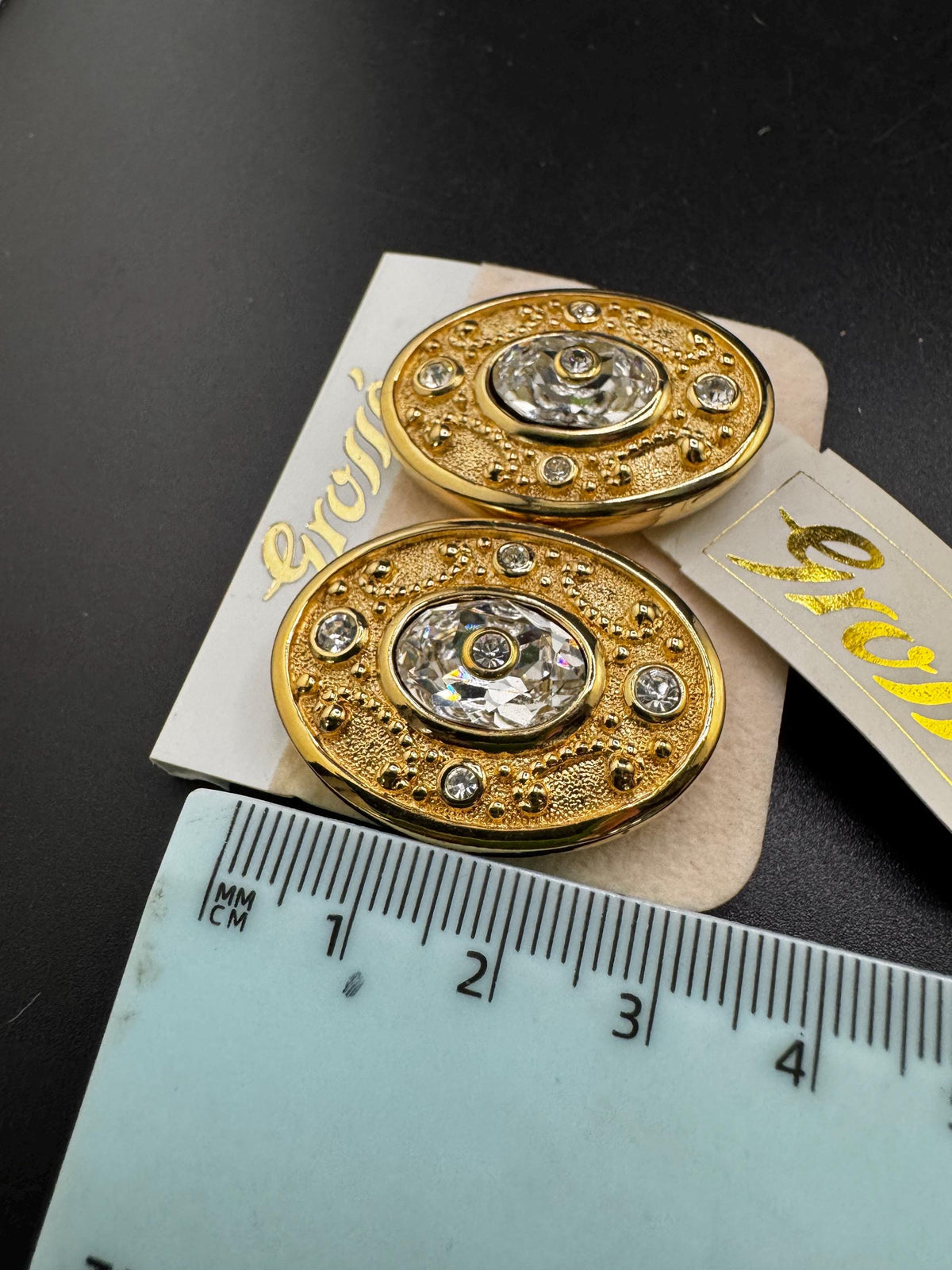 two ornate gold buttons with a central diamond, placed on a ruler for size comparison.