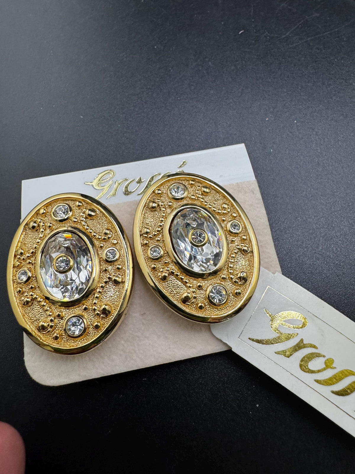 a pair of gold earrings with a clear gemstone in the center, placed on a white card with text.