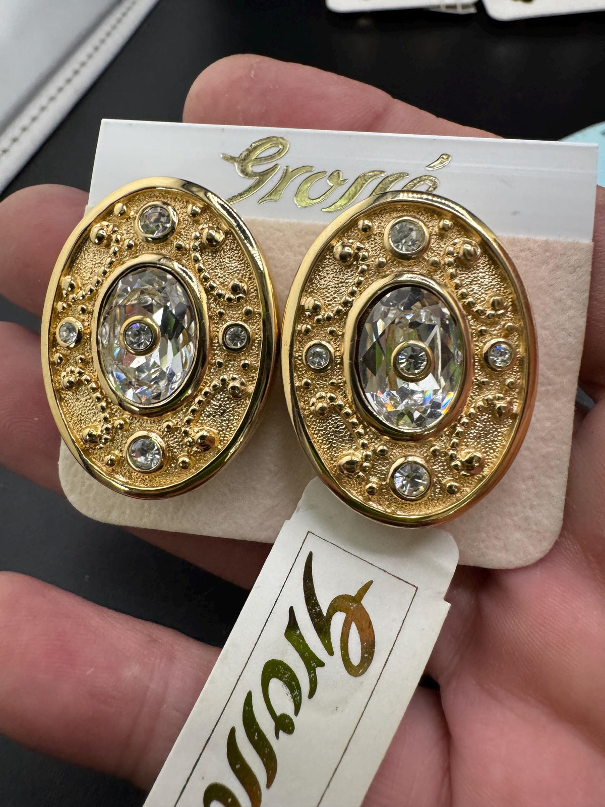 A pair of gold earrings with a large oval-shaped gemstone and smaller gemstones around it, displayed on a white card with text.