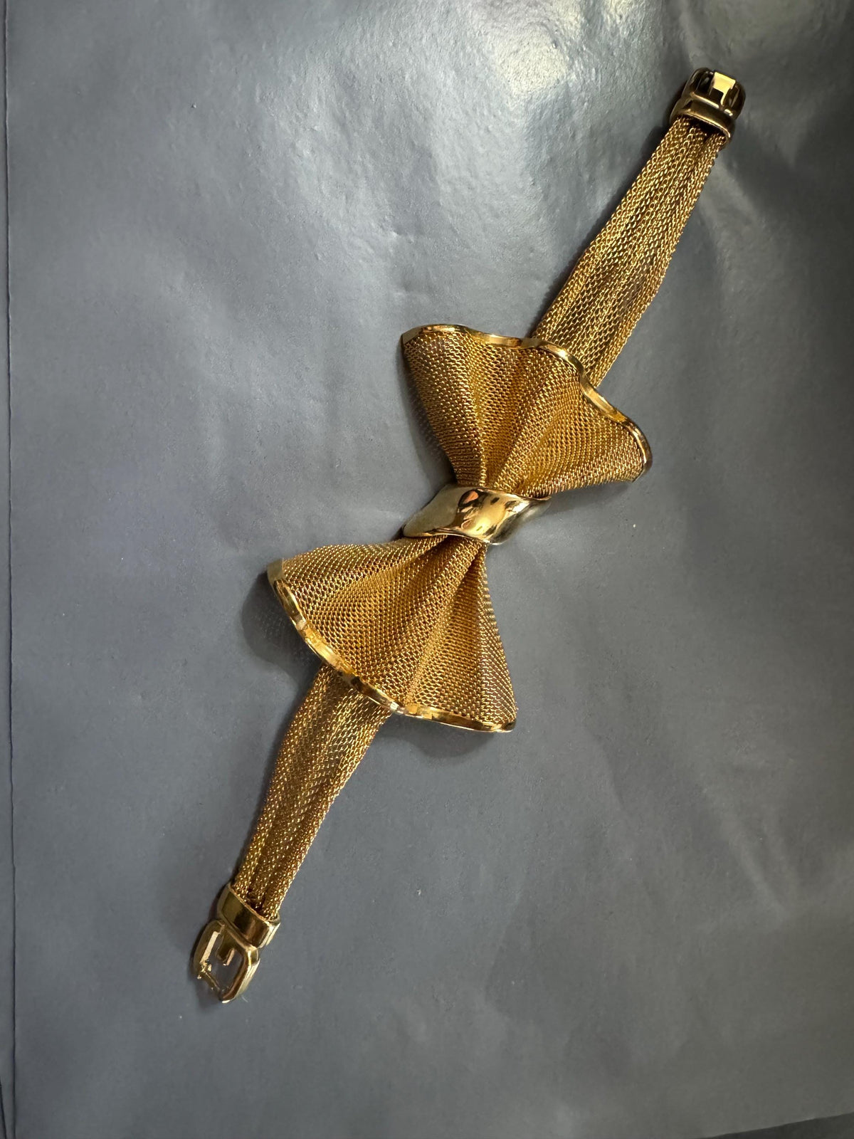 A gold-colored bow tie bracelet is shown against a gray background.