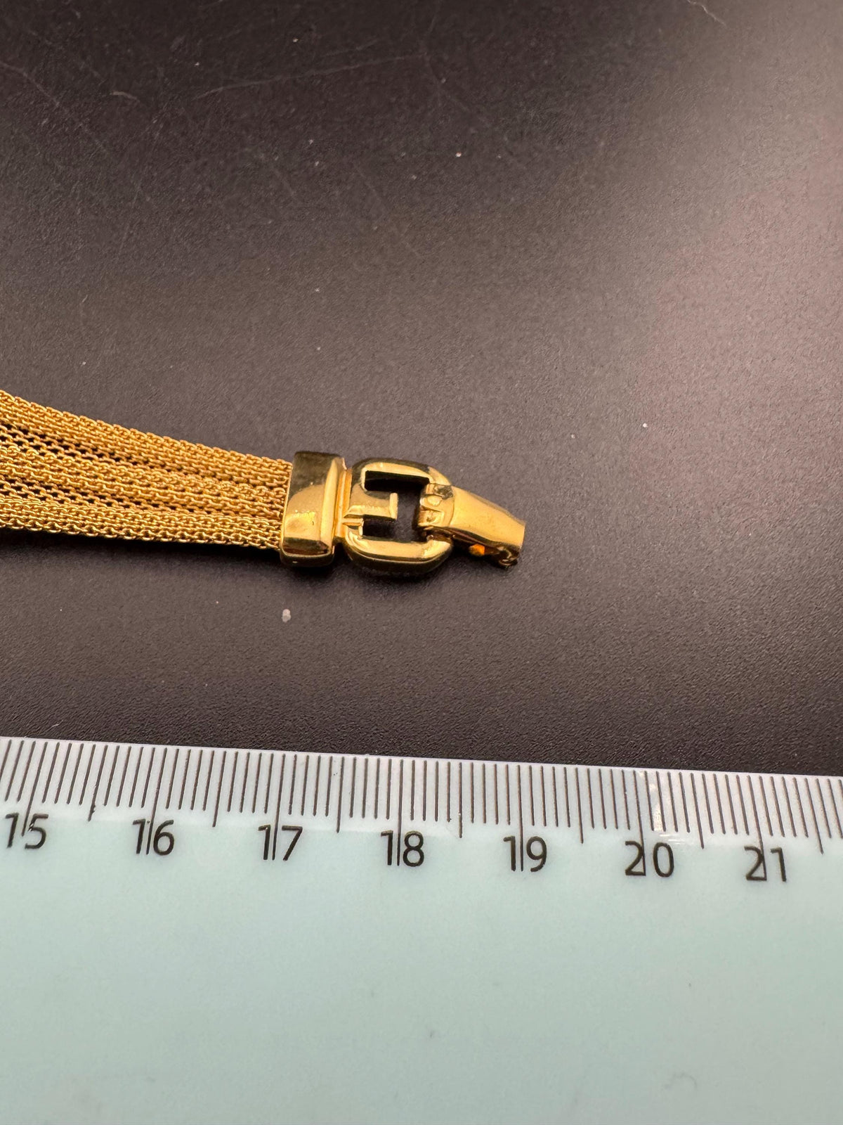 A gold-colored bracelet with a clasp is placed next to a ruler on a black surface.