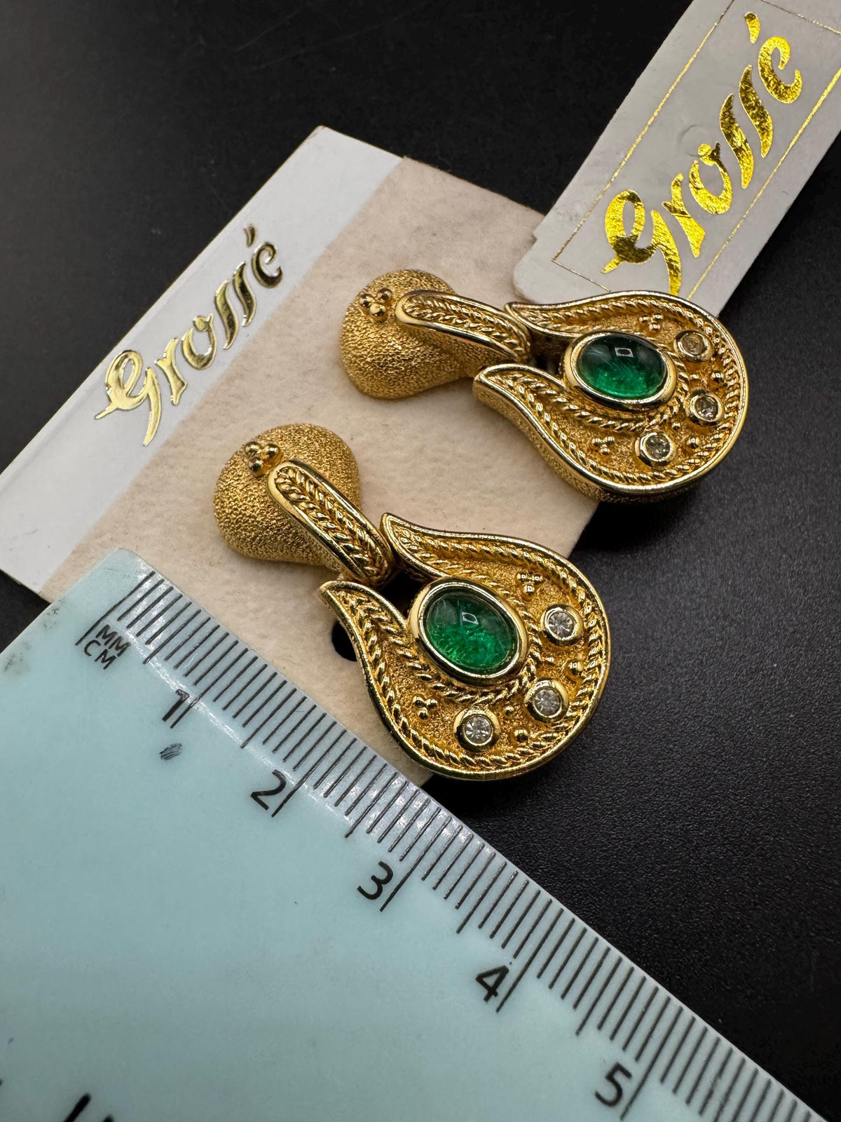a pair of ornate gold earrings with green gemstones, placed on a black surface next to a ruler for size comparison.