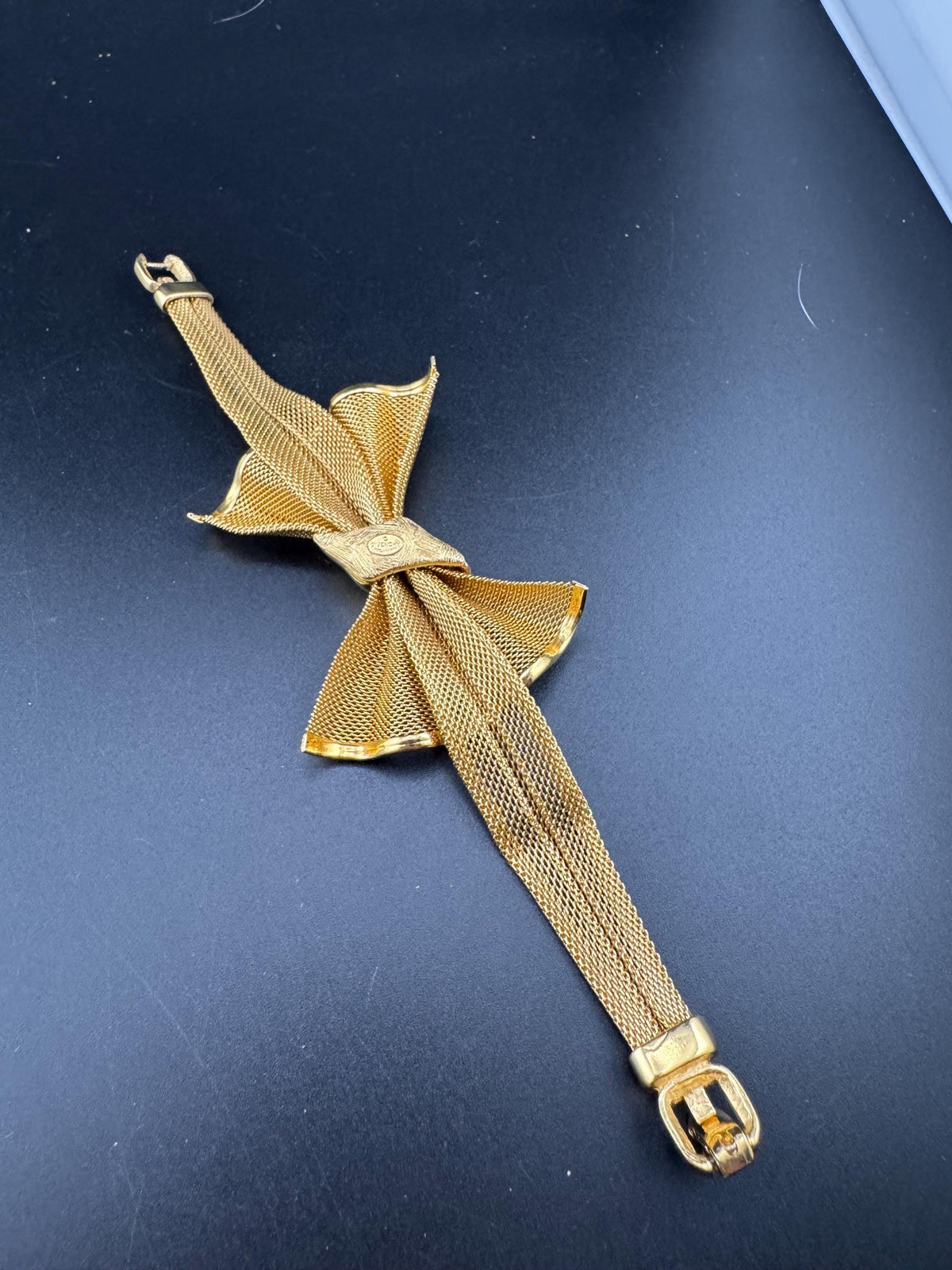 A gold-colored metal bracelet with a bow-shaped clasp is shown against a dark background.