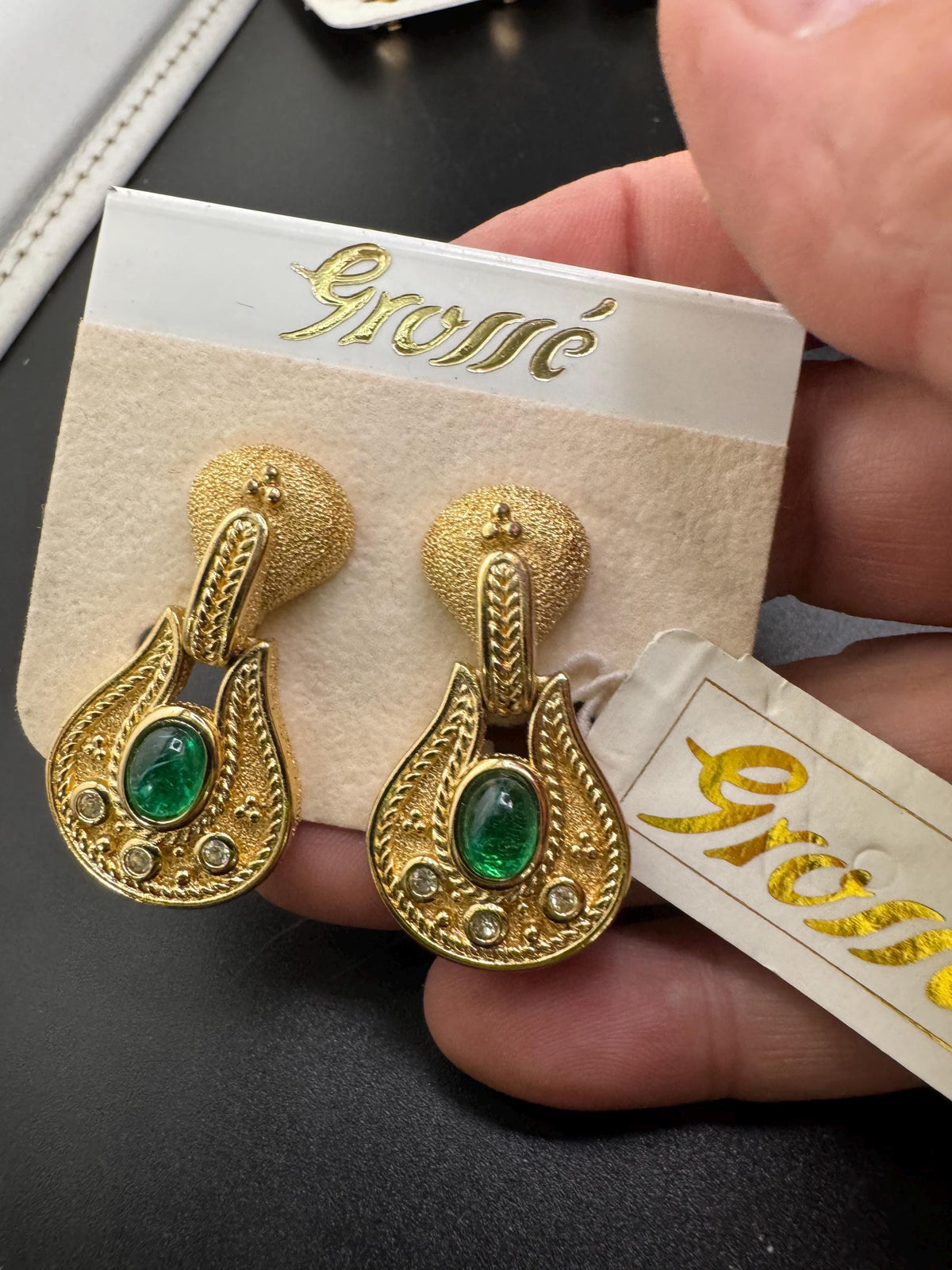 A person is holding a pair of gold earrings with green gemstones, and there is a tag attached to the earrings.