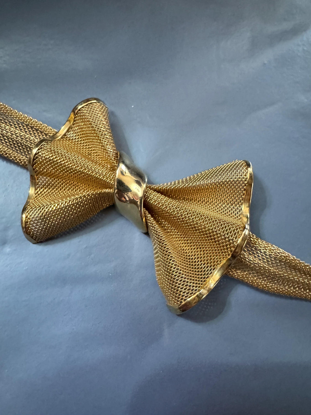 A gold-colored bow tie with a shiny, metallic appearance is displayed against a blue background.