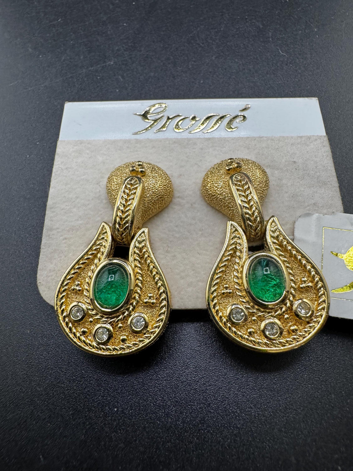 A pair of gold earrings with green gemstones and intricate designs, displayed on a white card with the word "Eme" written on it.